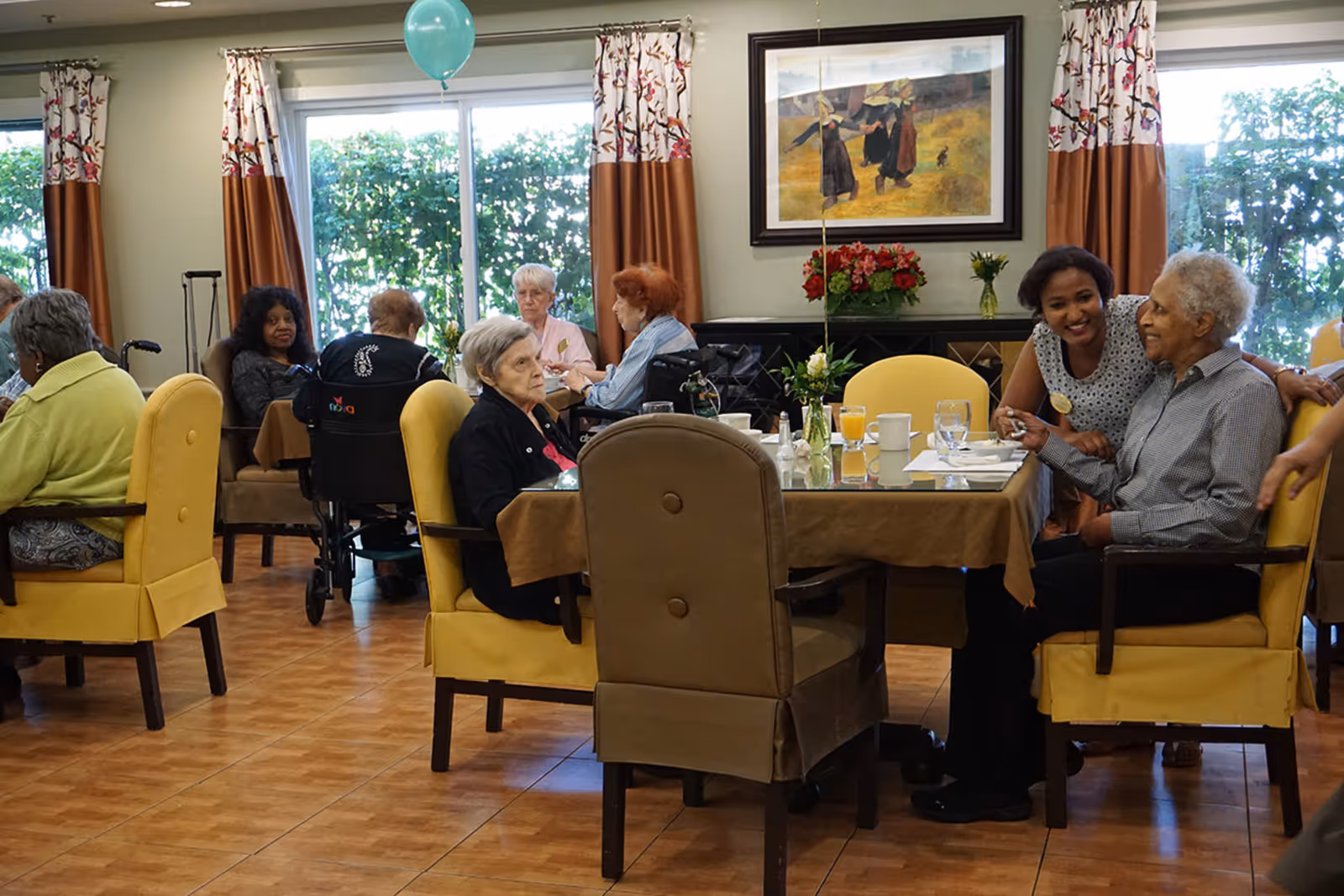 A group of elderly people sitting around tables in a dining room at a senior living facility, engaging in conversation and enjoying drinks. The room has large windows with curtains, a painting on the wall, and a balloon tied to one of the chairs.