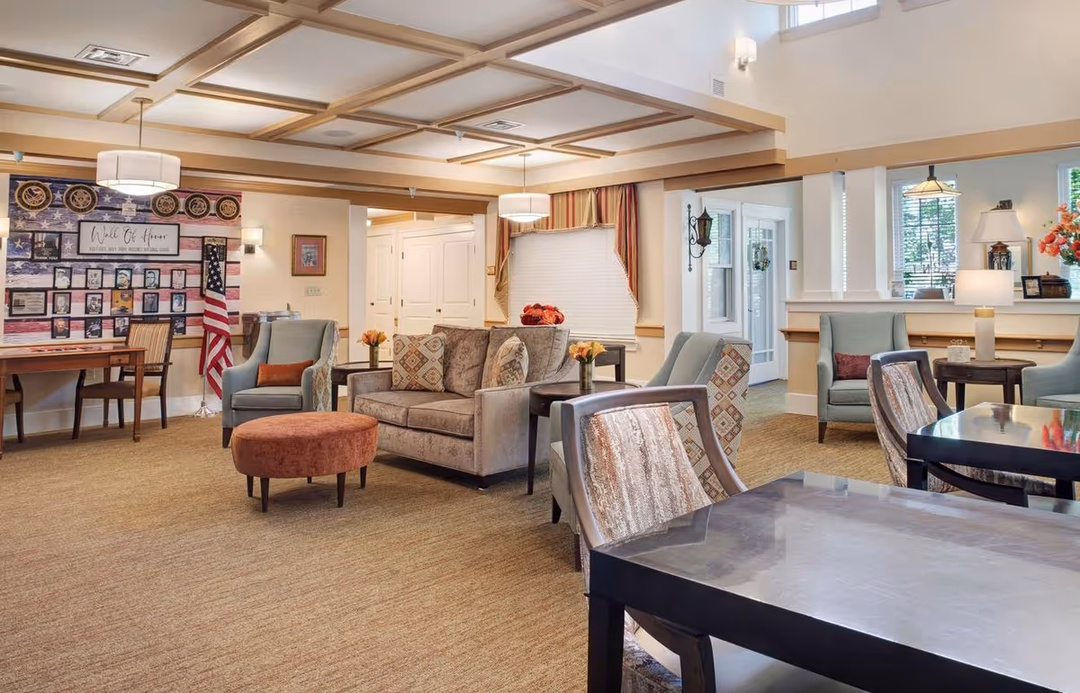 A bright communal lounge with sofas, armchairs, tables, an American flag and a bulletin board in a senior living facility.