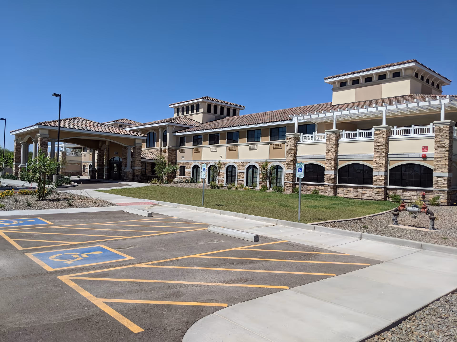 Exterior view of The Mission at Agua Fria Senior Living facility showing a large two-story building with beige walls, stone accents, and a tiled roof under a clear blue sky. In front of the building is a parking lot with marked handicapped parking spaces and landscaped areas with grass and small plants.
