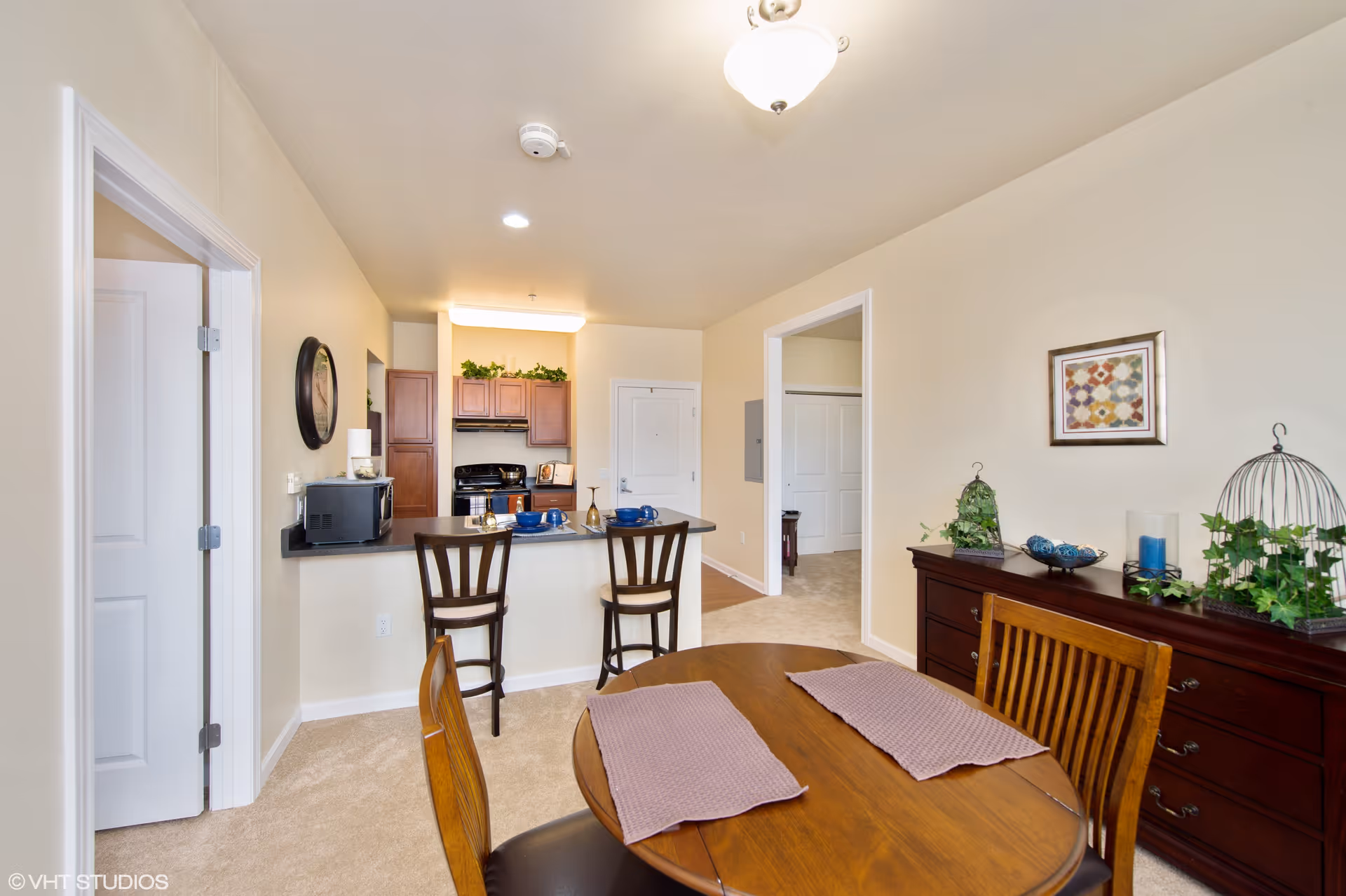 Interior view of a senior living facility showing a dining area with a wooden table and chairs, a sideboard with decorative items, and a kitchen with a breakfast bar and two high chairs. The walls are light-colored, and there are doors leading to other rooms.