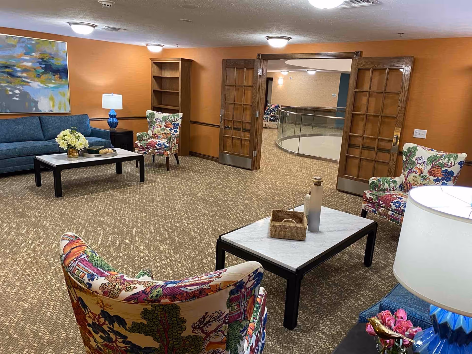 A cozy living room area with orange walls and patterned carpet. The room features a blue sofa, two floral-patterned armchairs, a coffee table with decorative items, a side table with a blue lamp, and a wooden bookshelf. Double wooden doors open to a hallway with a curved glass railing.