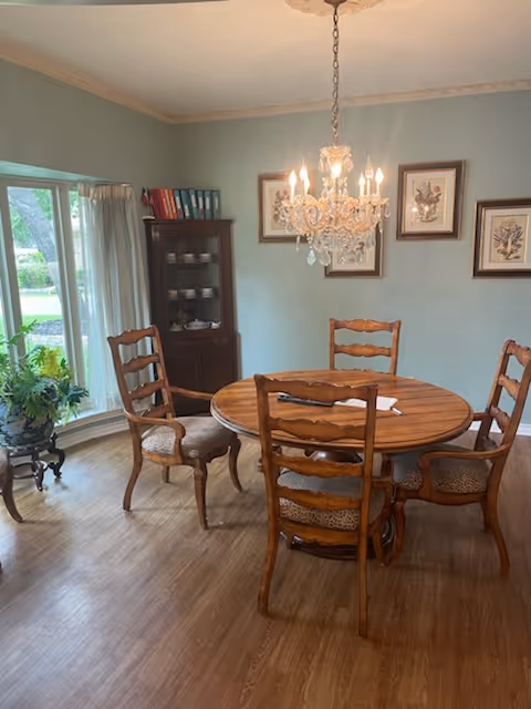 A dining room with a round wooden table surrounded by five wooden chairs with cushioned seats. A crystal chandelier hangs above the table. There is a wooden cabinet with shelves holding dishes and books against the wall. Three framed botanical prints are hung on the light blue wall. A large window with sheer white curtains lets in natural light, and a small plant sits on a decorative stand near the window. The floor is wooden.