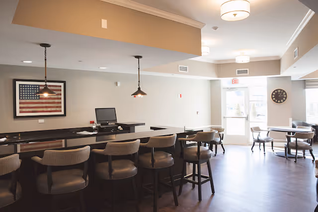 Communal dining and lounge area with a long bar counter, multiple stools, tables, pendant lights, and framed American flag art.