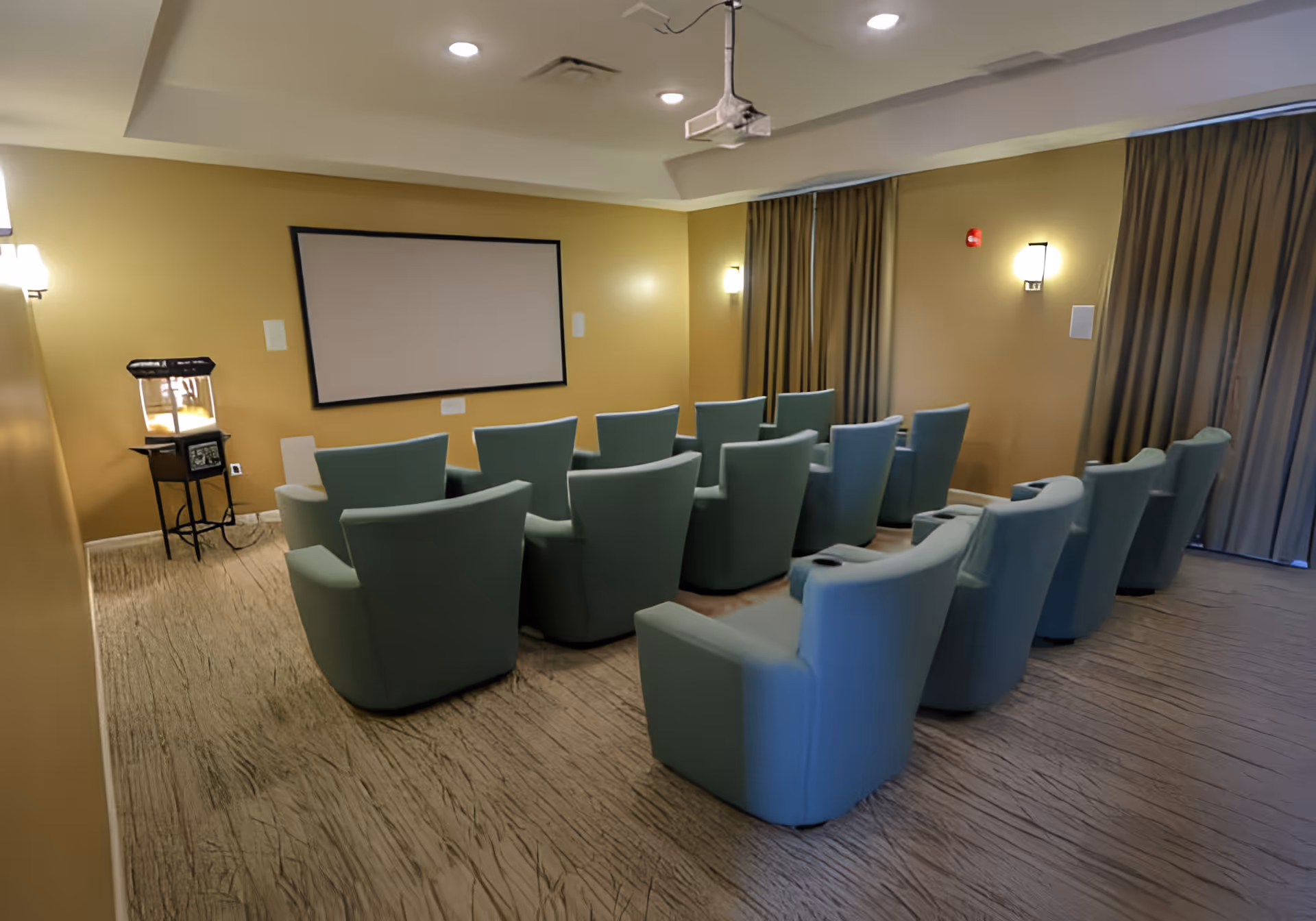 A small theater room with two rows of light blue cushioned chairs facing a blank projector screen on a yellow wall. There is a popcorn machine on a stand to the left side of the room and curtains covering windows on the right side. The room has soft lighting from wall sconces and a ceiling-mounted projector.