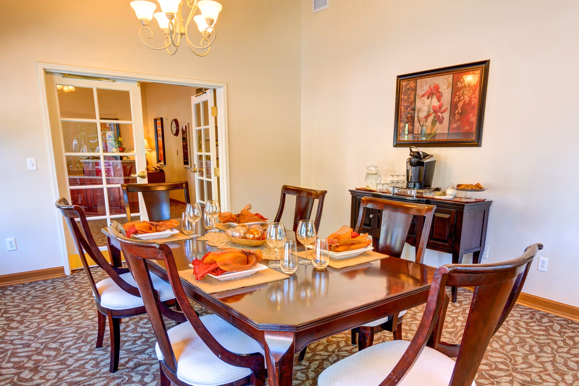 A dining room with a polished wooden table set for six people with orange and red napkins, glassware, and placemats. There are six wooden chairs with white cushions around the table. In the background, there is a wooden sideboard with a coffee maker, glasses, and a plate of pastries. A framed floral painting hangs on the wall above the sideboard. The room has beige walls, patterned carpet, and a chandelier overhead. French doors lead to another room.