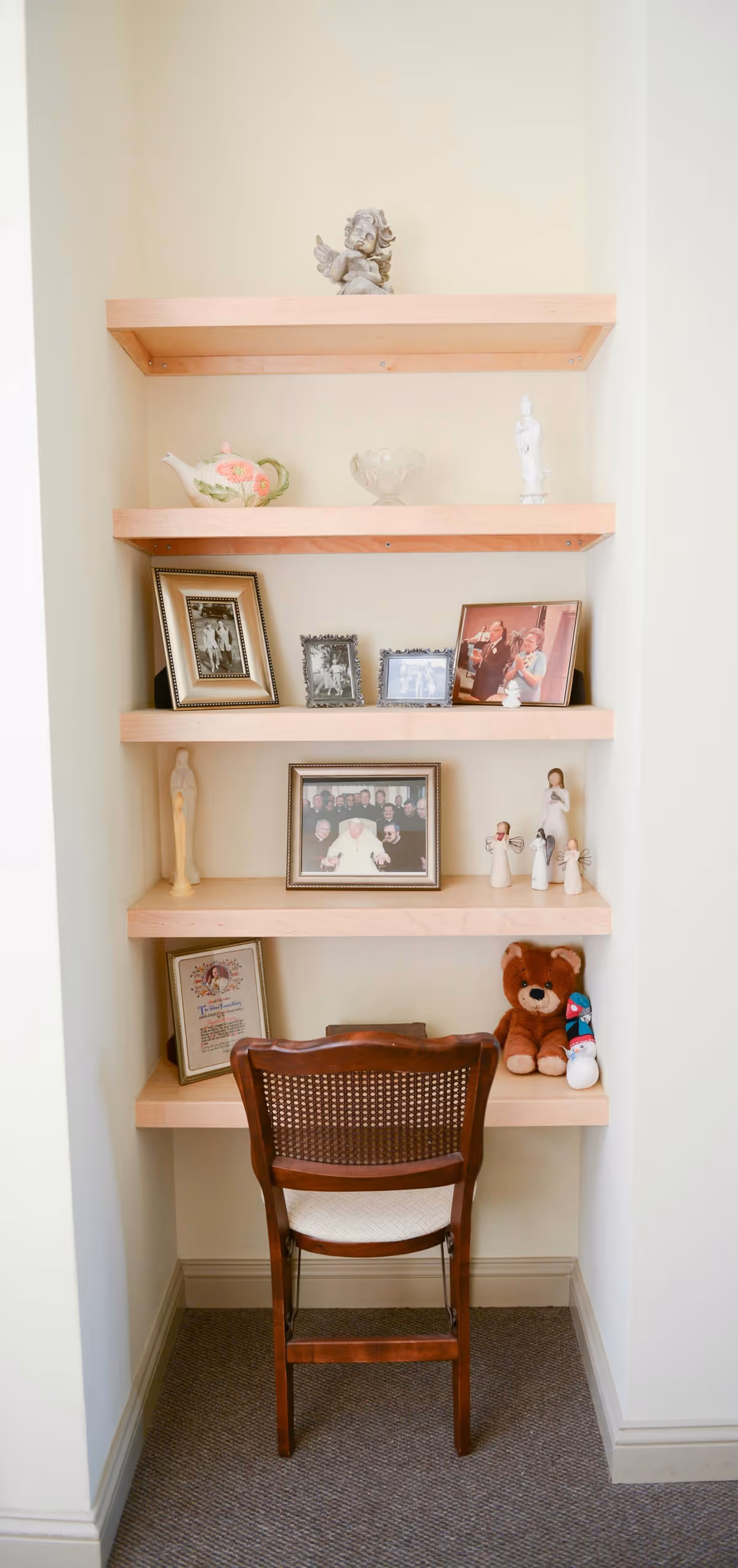 A small nook with four wooden shelves mounted on the wall, displaying various decorative items including framed photographs, figurines, a teapot, a teddy bear, and a certificate. A wooden chair with a woven backrest is placed in front of the shelves.