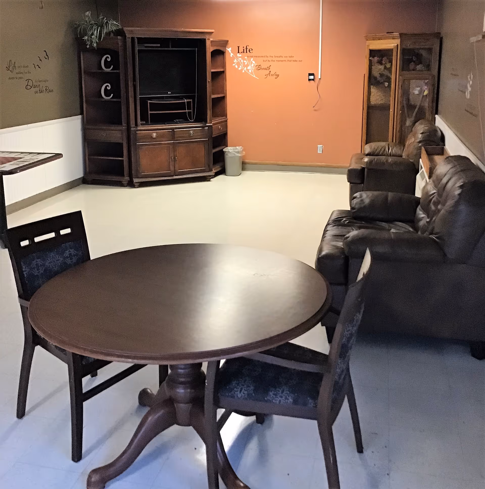 A common living area with a round wooden table and chairs in the foreground, leather recliners along the right wall, and an entertainment cabinet against a warm-colored wall.
