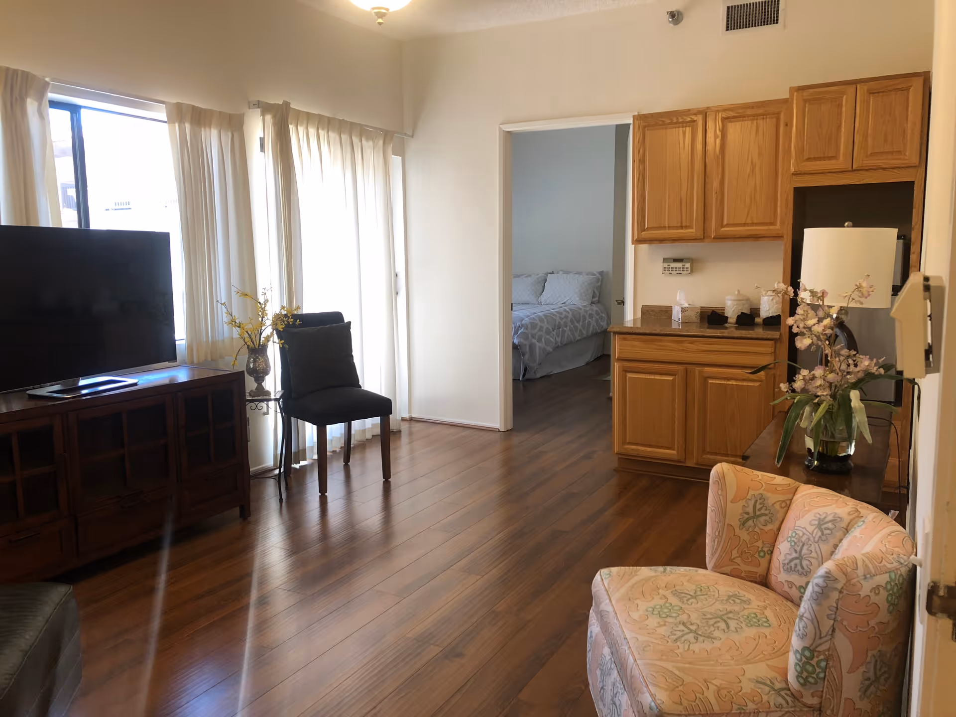 Interior view of a senior living facility room with wooden flooring, a large flat-screen TV on a wooden cabinet, a black chair next to a window with white curtains, a floral upholstered armchair, and wooden cabinets with a countertop. A doorway leads to a bedroom with a bed covered in gray and white bedding.