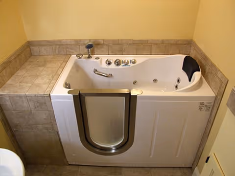 A walk-in bathtub with a side door, built-in seat, and multiple control knobs, surrounded by beige tiled walls and floor in a bathroom setting.