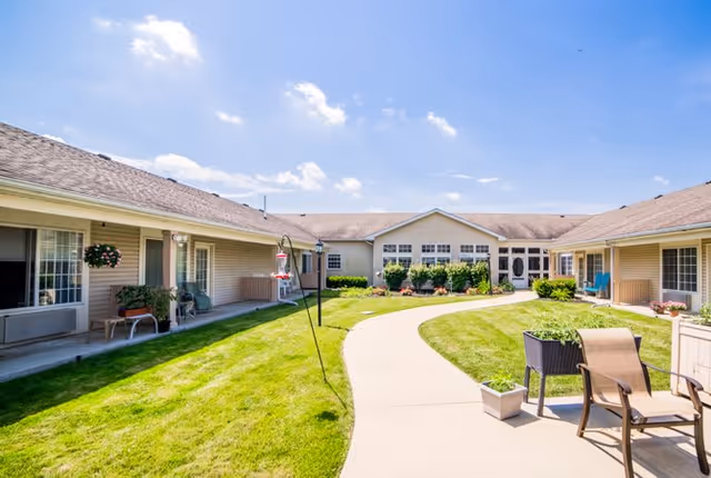 Outdoor courtyard area of Kokomo Place senior living facility with a curved concrete walkway, green grass, patio chairs, potted plants, and a clear blue sky with some clouds.