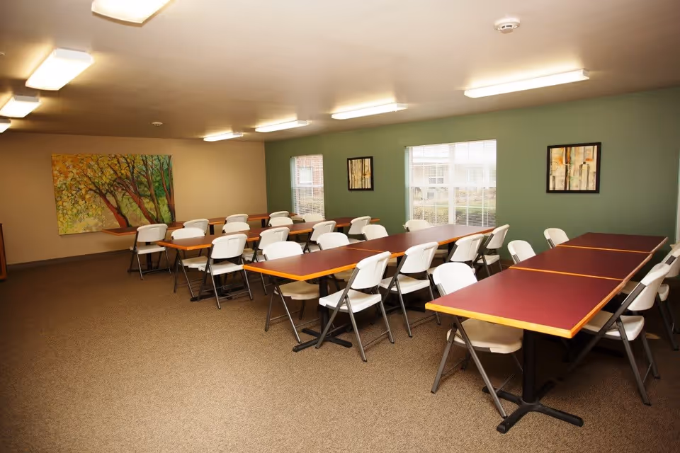 A dining room with several rectangular tables arranged in rows, surrounded by white folding chairs. The room has green and beige walls with three framed artworks, two windows letting in natural light, and ceiling lights providing illumination.
