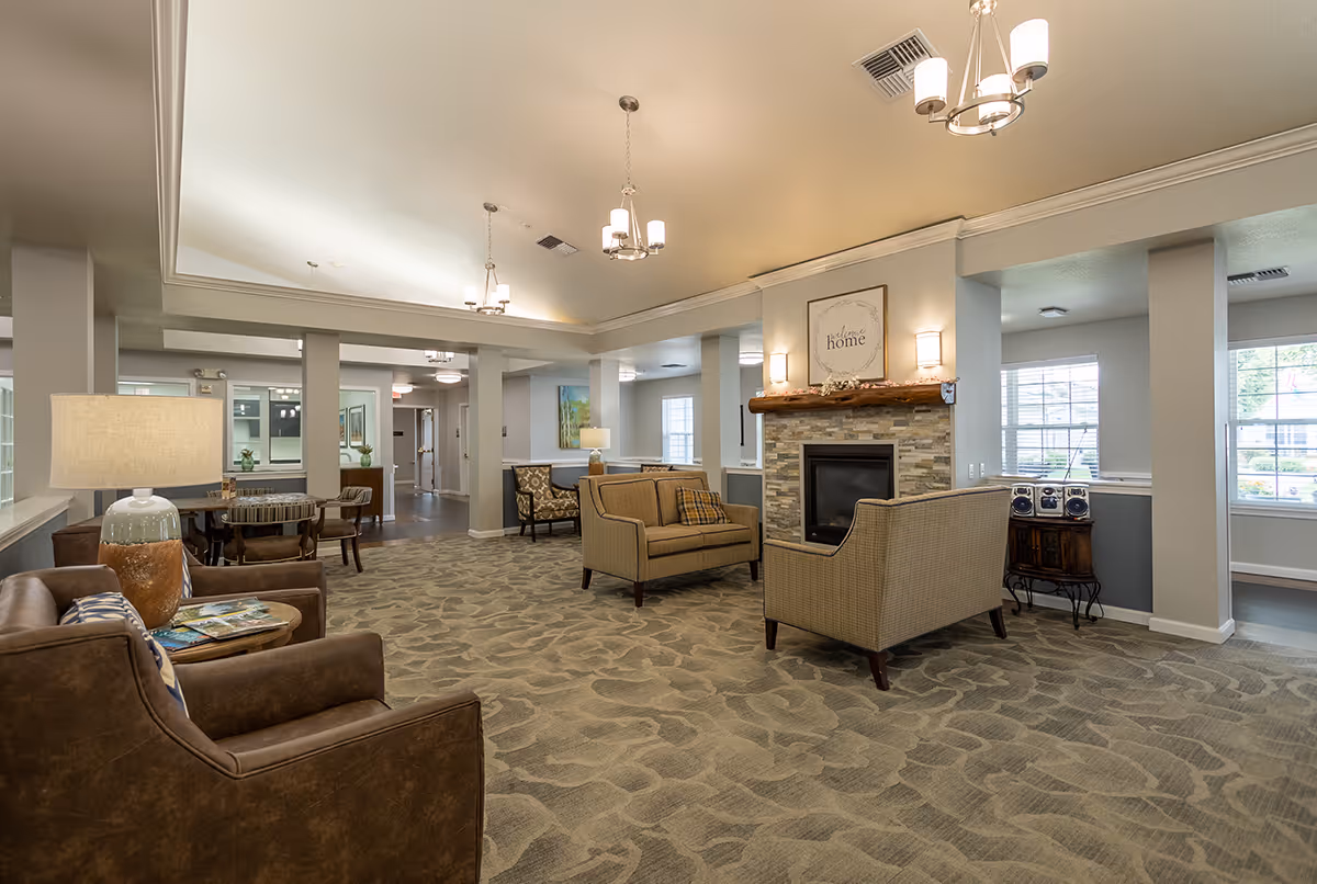 Spacious, well-lit senior living common area with armchairs, sofas, a fireplace, and a small dining table.