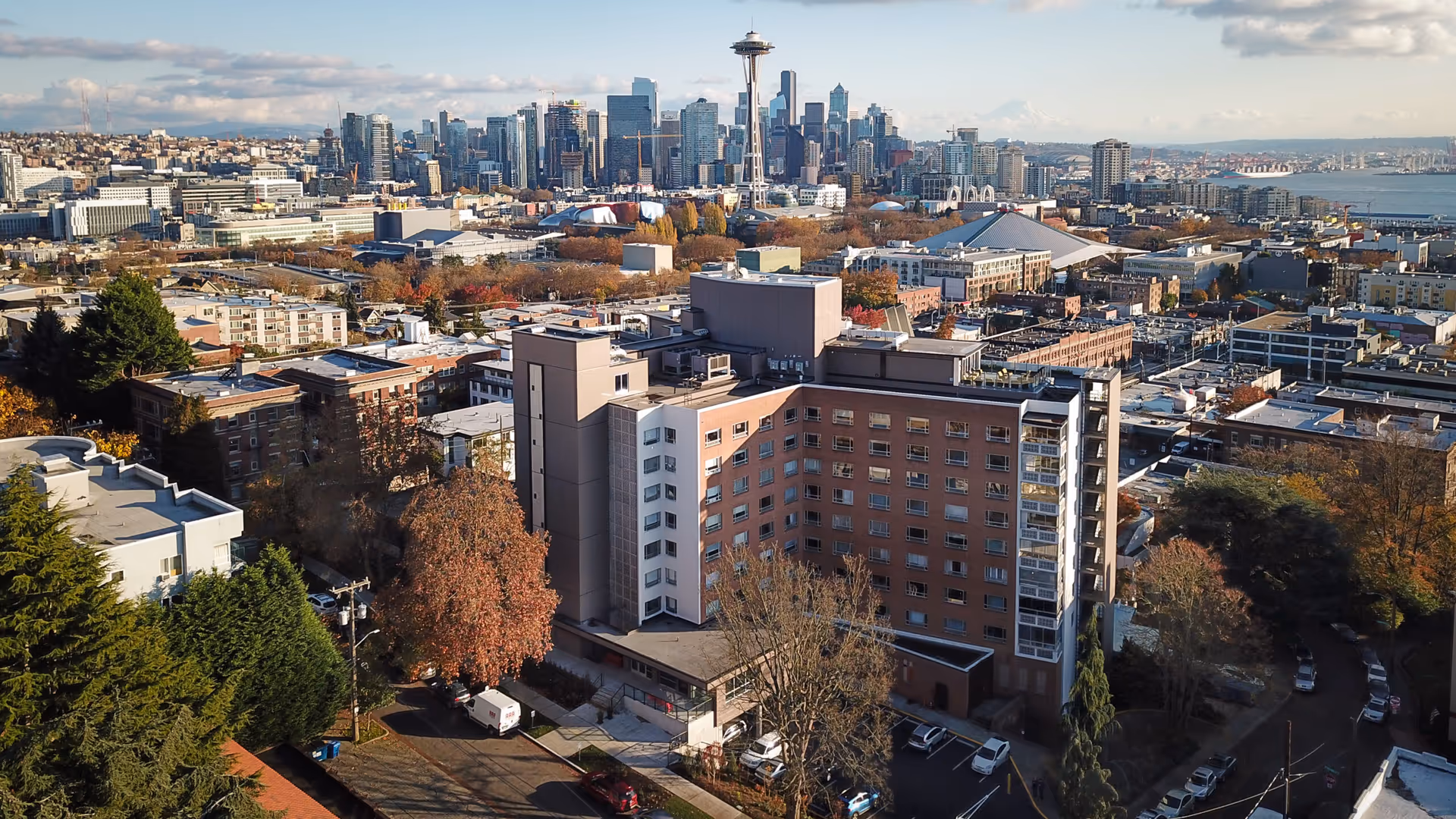Aerial view of a large multi-story building surrounded by trees and smaller buildings with a city skyline in the background, including the iconic Space Needle and Mount Rainier in the distance under a partly cloudy sky.