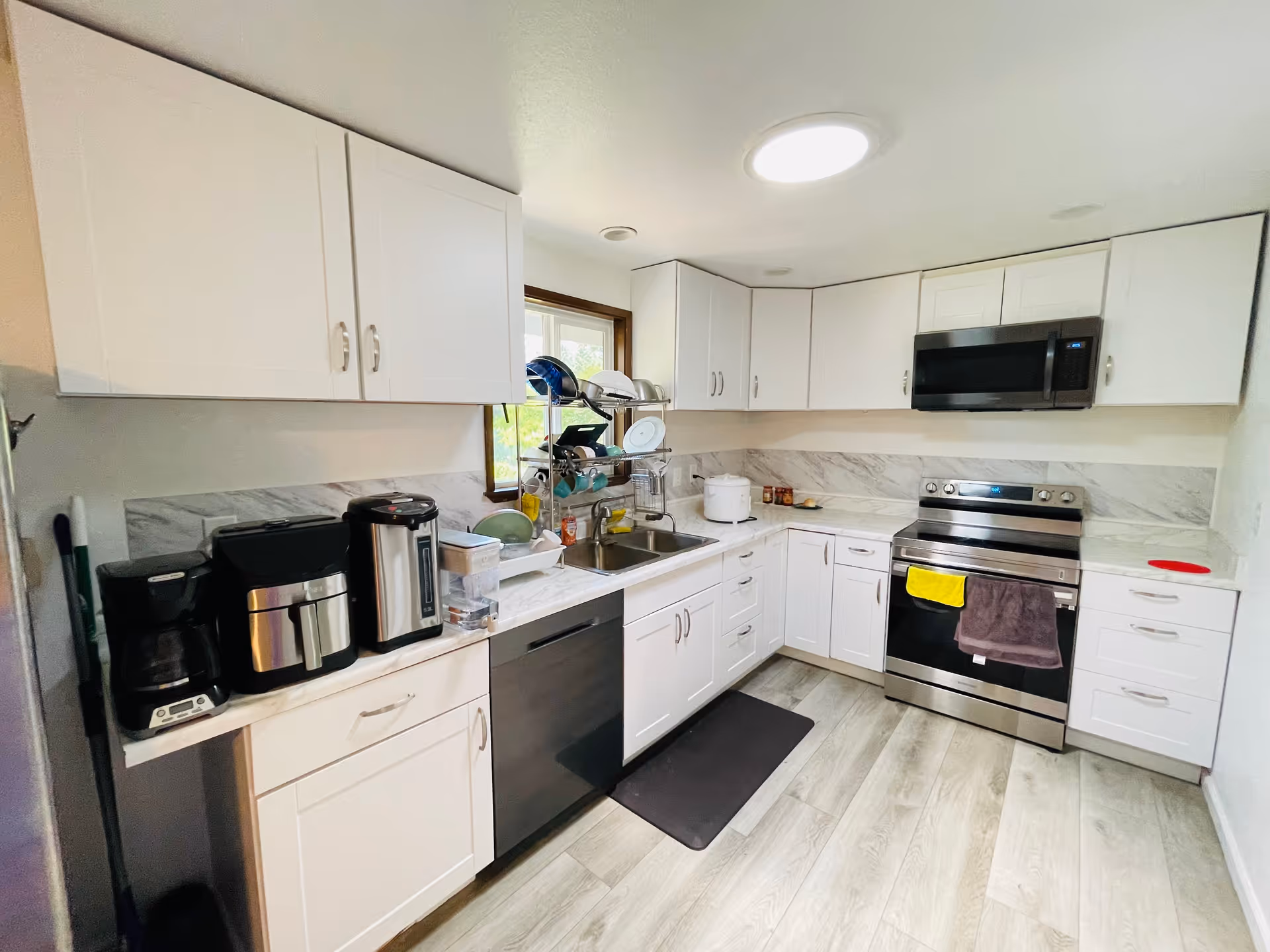 A modern kitchen with white cabinets and drawers, a stainless steel electric stove with a microwave above it, a dishwasher, a double sink with a dish rack, and various kitchen appliances including a coffee maker and an air fryer. The kitchen has light wood flooring and a window above the sink.