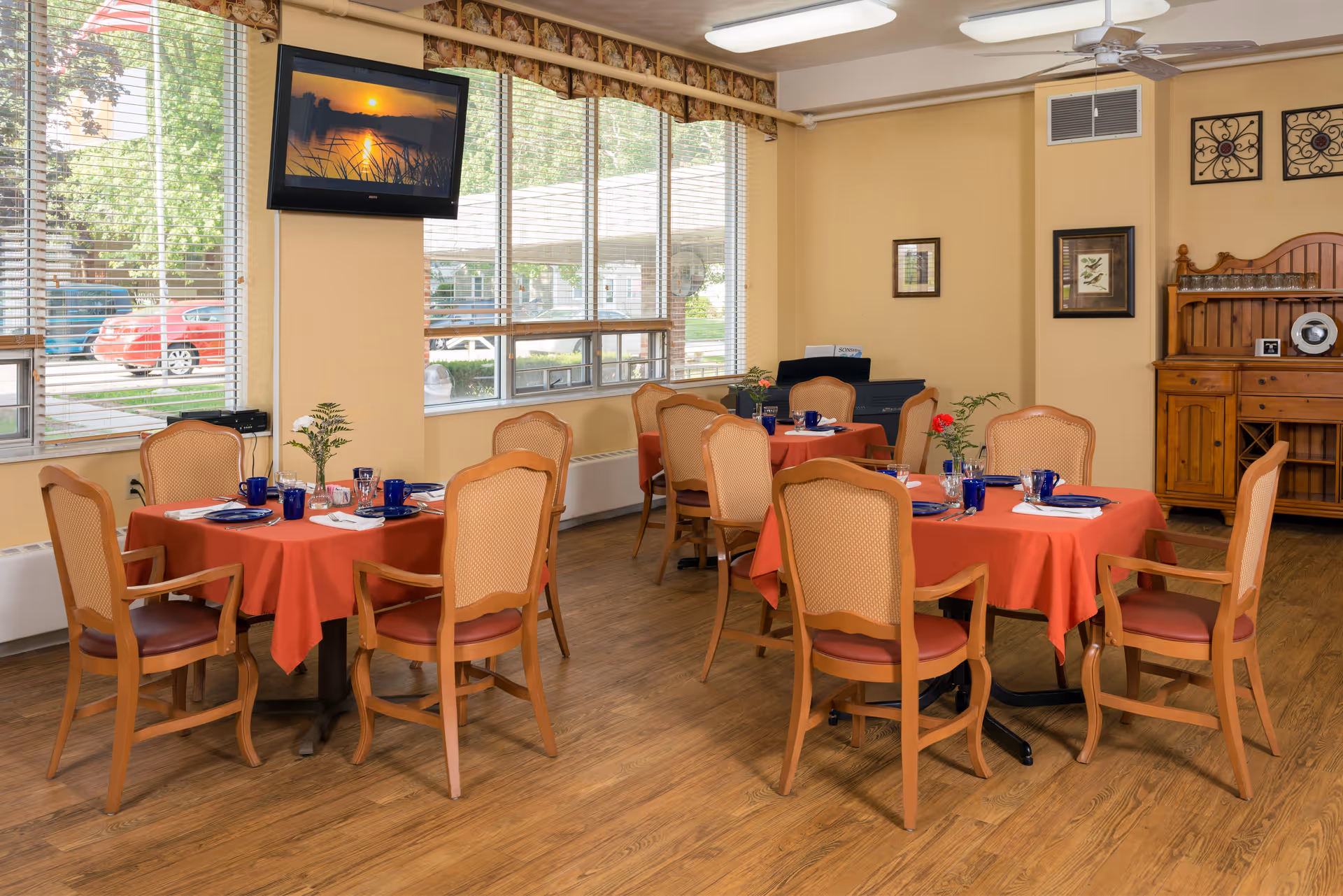 A dining room with several round tables covered with red tablecloths, each set with blue glasses, white napkins, and small flower arrangements. Wooden chairs with cushioned seats surround the tables. Large windows with blinds let in natural light, and a flat-screen TV is mounted on the wall showing a sunset scene. The room has wooden flooring and beige walls with framed artwork and a wooden cabinet in the corner.
