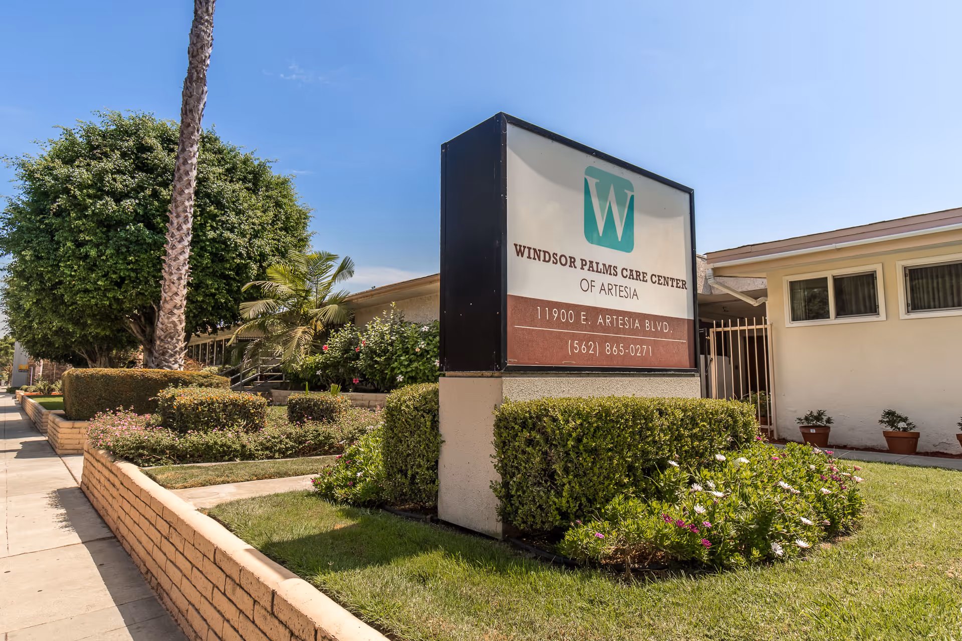 Outdoor view of Windsor Palms Care Center of Artesia sign with surrounding greenery, bushes, flowers, and a building in the background under a clear blue sky.