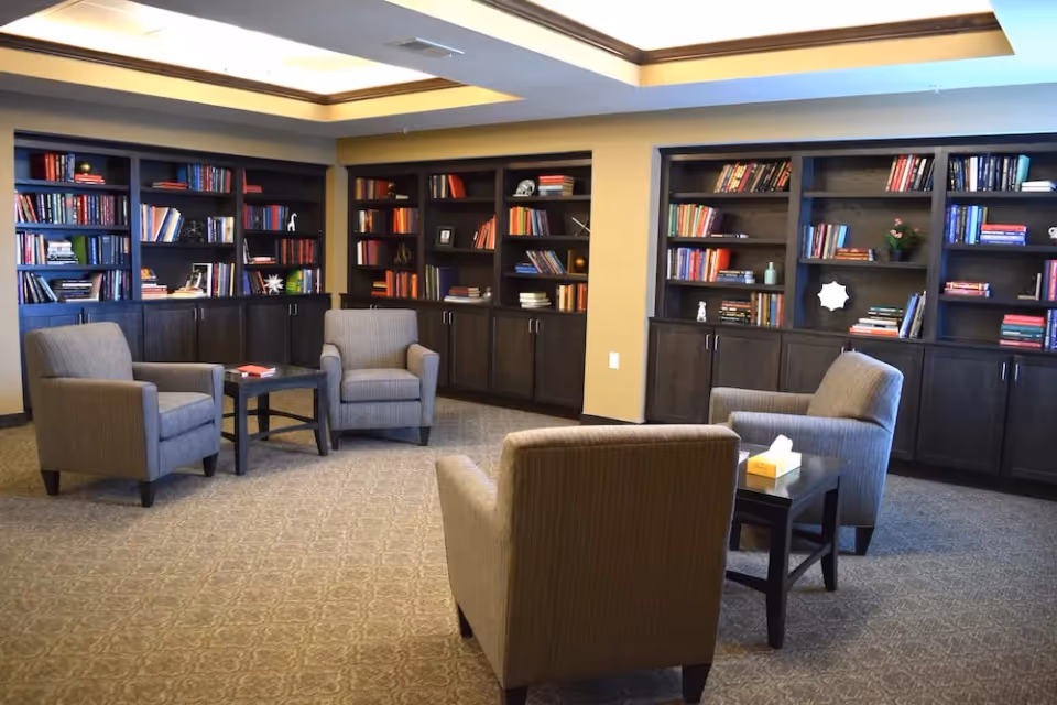 A cozy library-style sitting room with four upholstered armchairs around small tables and wall-to-wall bookshelves.