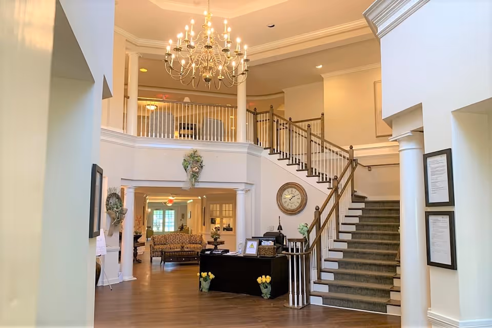 Interior view of a senior living facility lobby with a grand staircase, chandelier, and seating area. The space features white walls, wooden flooring, decorative columns, and framed documents on the walls. A reception desk is positioned near the staircase with flower arrangements nearby.