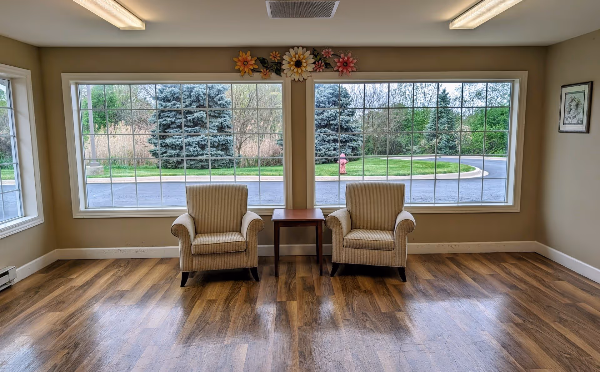 A bright room with two beige armchairs separated by a small wooden table, positioned in front of large windows showing green trees and a fire hydrant outside. The room has wooden flooring, beige walls, and decorative flower wall art above the windows.