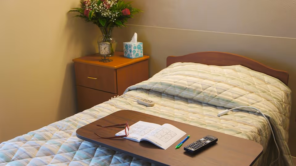 A neatly made single bed in a nursing facility room with a bedside table, flowers and tissue box, and an overbed tray holding glasses, a book, pens, and remote controls.