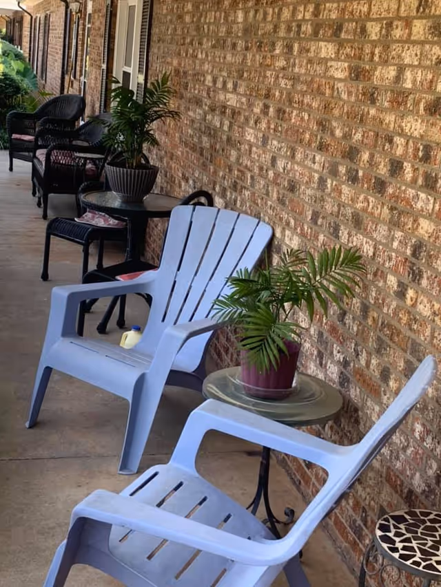 A covered outdoor walkway with a brick wall on one side and several chairs and small tables arranged along the path. There are two light blue plastic chairs in the foreground with a small round glass table holding a potted plant between them. Further down the walkway, there are black wicker chairs with cushions and more potted plants on tables.