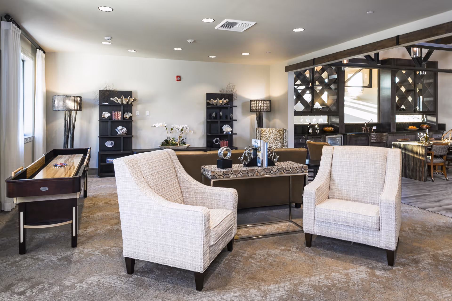 Bright communal lounge with two upholstered armchairs facing a sofa, a shuffleboard table to the left, and a bar/dining area in the background.