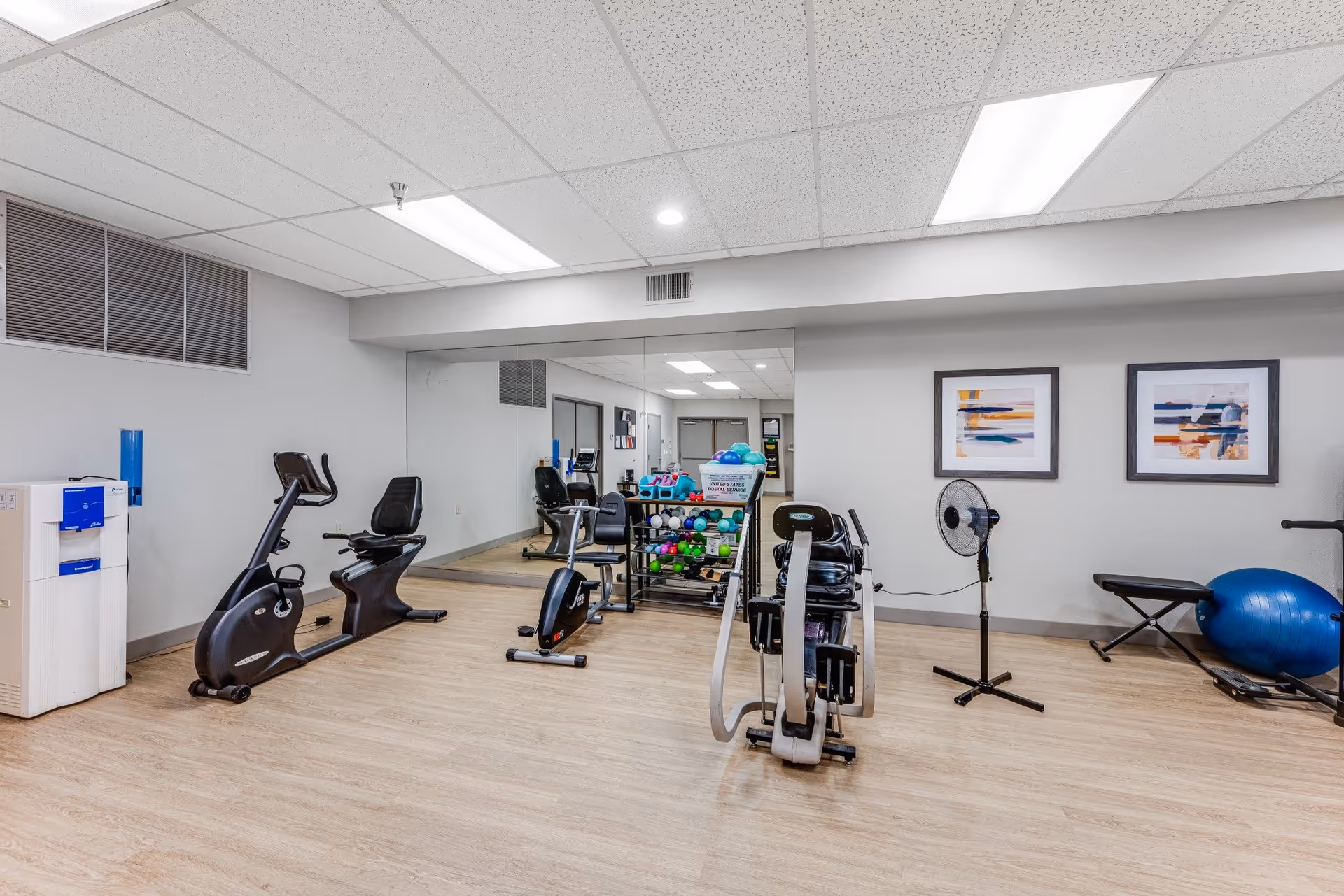 A fitness room with exercise equipment including two stationary bikes, a rowing machine, a rack of colorful dumbbells, a blue exercise ball, a bench, a standing fan, and two abstract paintings on the wall. The room has wood flooring, white walls, a drop ceiling with fluorescent lights, and a large mirror covering one wall.