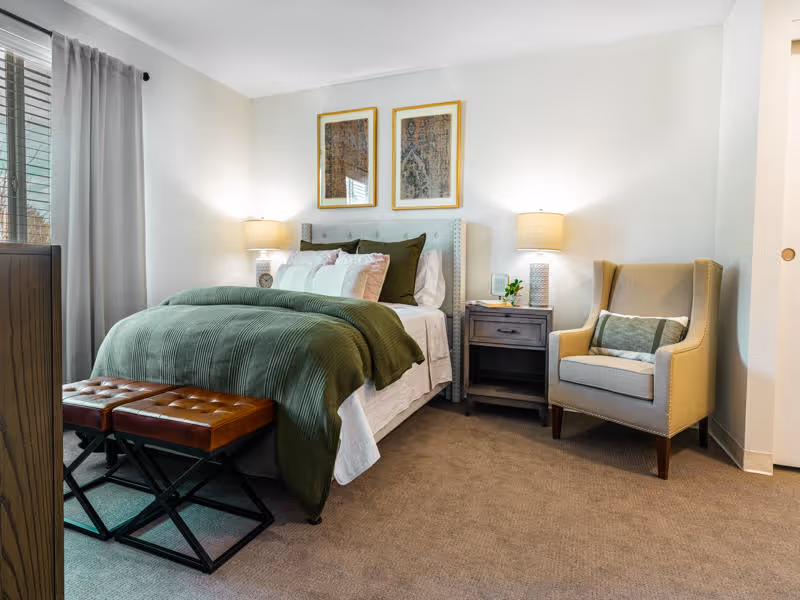 A cozy bedroom with a neatly made bed featuring a green blanket and multiple pillows. Two framed artworks hang above the bed. There are two bedside tables with lamps on each side of the bed. A beige armchair with a decorative pillow is placed to the right of the bed. Two brown cushioned benches are positioned at the foot of the bed. A window with gray curtains is visible on the left side.