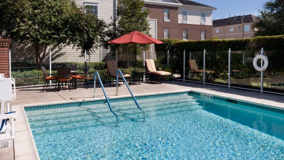 Outdoor swimming pool with clear blue water, surrounded by a concrete deck with lounge chairs, a table with chairs under a red umbrella, and a safety fence with a life preserver. Residential buildings and greenery are visible in the background.