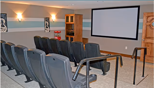 A small theater room with two rows of black cushioned seats facing a large blank projector screen mounted on a beige wall. The room has soft lighting from wall sconces, two framed posters on the wall, a wooden cabinet with electronic equipment, and a red popcorn machine in the corner. There are handrails along the aisle between the seats.