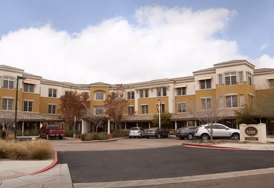 Exterior view of a three-story senior living facility building with beige and yellow walls, multiple windows, and a tiled roof. Several cars are parked in front of the building, and there are some trees and shrubs around the entrance. A sign near the entrance reads 'Sunrise of Scottsdale.'