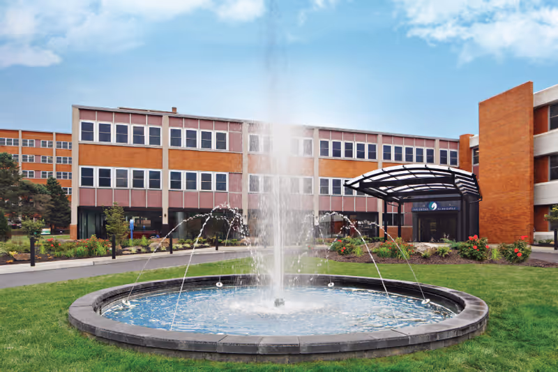 Exterior view of a multi-story brick building with many windows, featuring a circular water fountain in the foreground surrounded by green grass and landscaped plants under a partly cloudy sky.