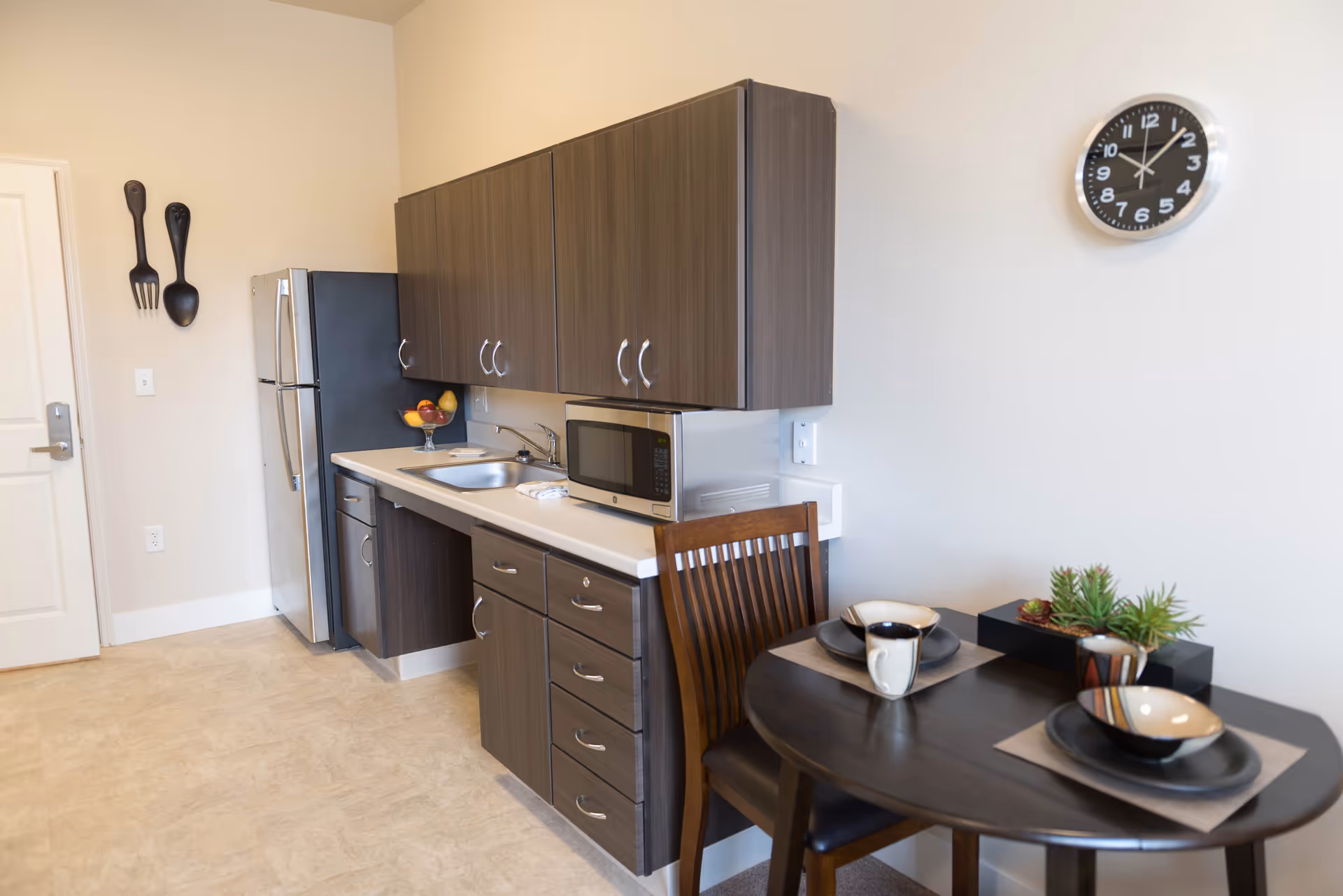 A small kitchen area with dark wood cabinets, a stainless steel refrigerator, a microwave on the counter, and a sink. A round dark wood table with two place settings and a small plant centerpiece is positioned near the kitchen. A black wall clock is mounted on the white wall above the table. Decorative oversized fork and spoon hang on the wall near the door.