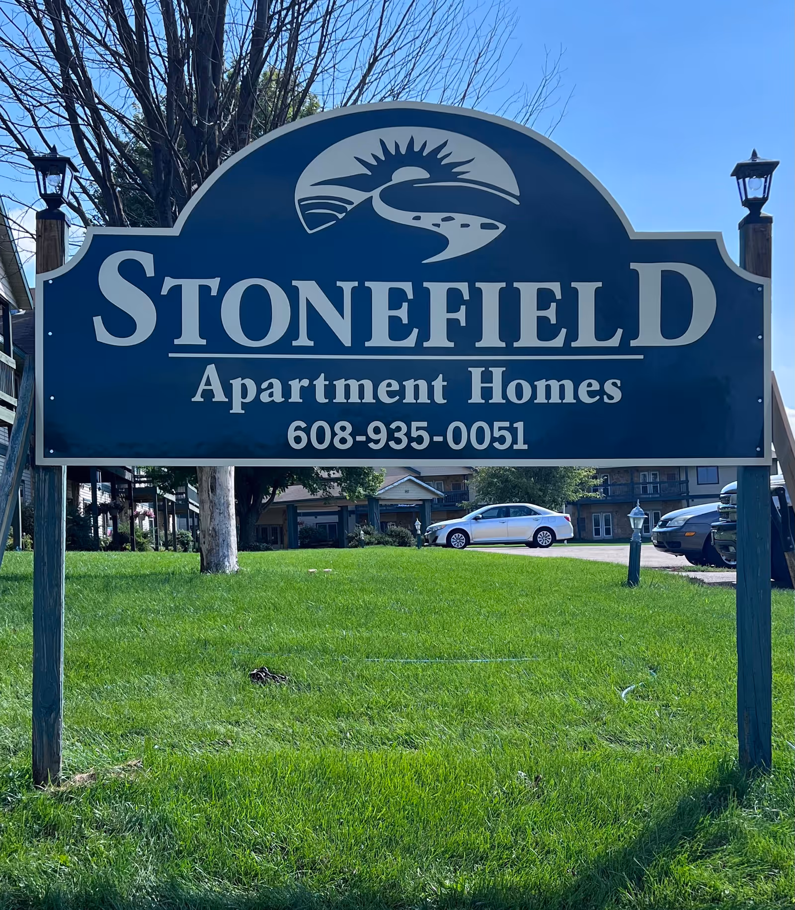 Large blue Stonefield Apartment Homes sign on a grassy lawn with apartment buildings and parked cars in the background.