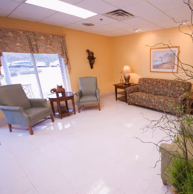 A cozy sitting area with two green armchairs and a floral patterned sofa arranged around wooden side tables. A lamp on one table provides warm lighting, and a framed picture hangs on the yellow wall. A large window with floral curtains lets in natural light, and a decorative plant is visible in the foreground.