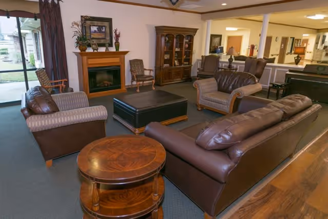 A cozy living room area with brown leather sofas and armchairs arranged around a large black ottoman. There is a wooden side table in the foreground, a fireplace with decorative items on the mantel, a wooden cabinet, and a window with curtains letting in natural light. The room has carpeted flooring and an open layout leading to other parts of the facility.