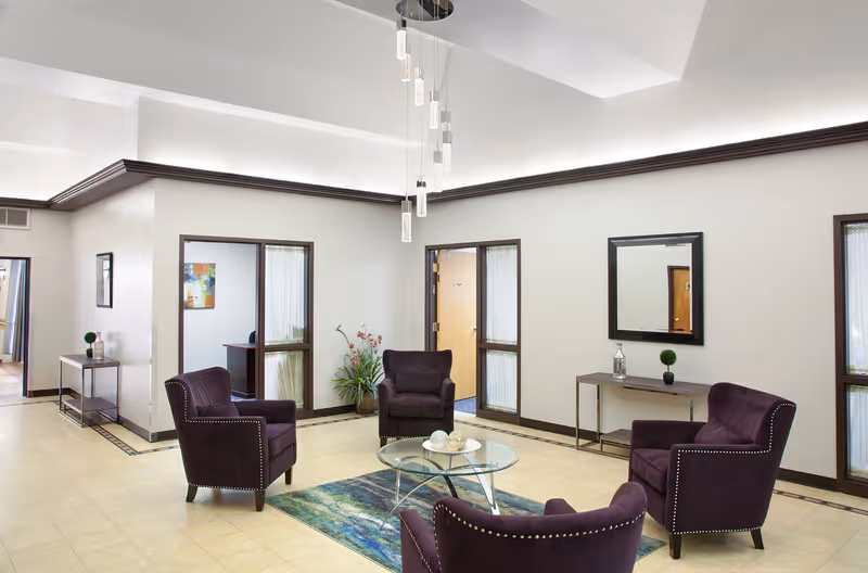 Bright communal sitting area with four purple armchairs arranged around a glass coffee table under a modern chandelier.