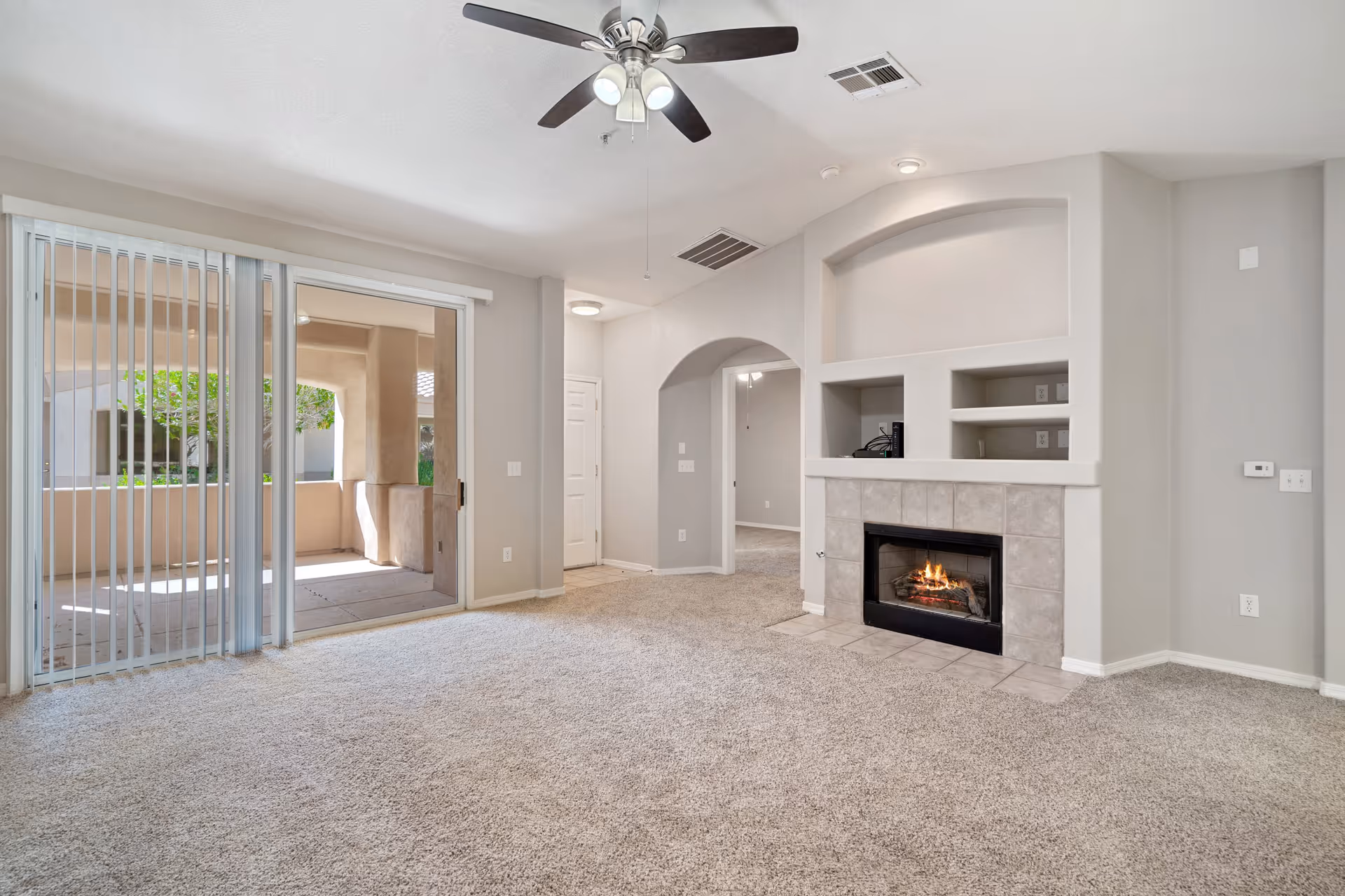 A bright living room with beige carpet, a ceiling fan with lights, a built-in fireplace with tiled surround, and built-in shelves above the fireplace. There is a sliding glass door with vertical blinds leading to a covered outdoor patio area with beige walls and greenery visible outside. An arched doorway leads to another room.