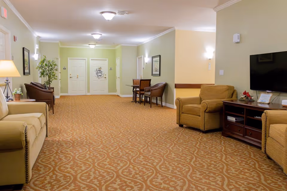 A well-lit interior common area with patterned carpet, beige walls, and ceiling lights. The space features comfortable seating including armchairs and a sofa, a small table with chairs, a flat-screen TV on a wooden stand, and decorative plants and framed pictures on the walls. There are multiple white doors at the far end, one with a 'home sweet home' sign.