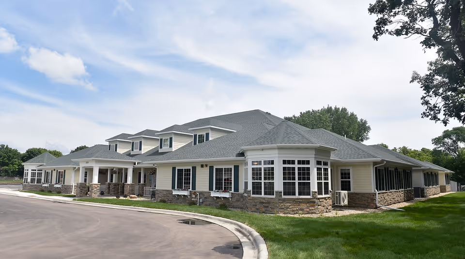 Front exterior of a low-rise residential building with stone accents, multiple windows, and a curved driveway under a partly cloudy sky.