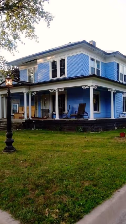 A two-story blue house with white trim and a wraparound porch featuring several chairs. The house is surrounded by a green lawn and a black street lamp is visible in the foreground.