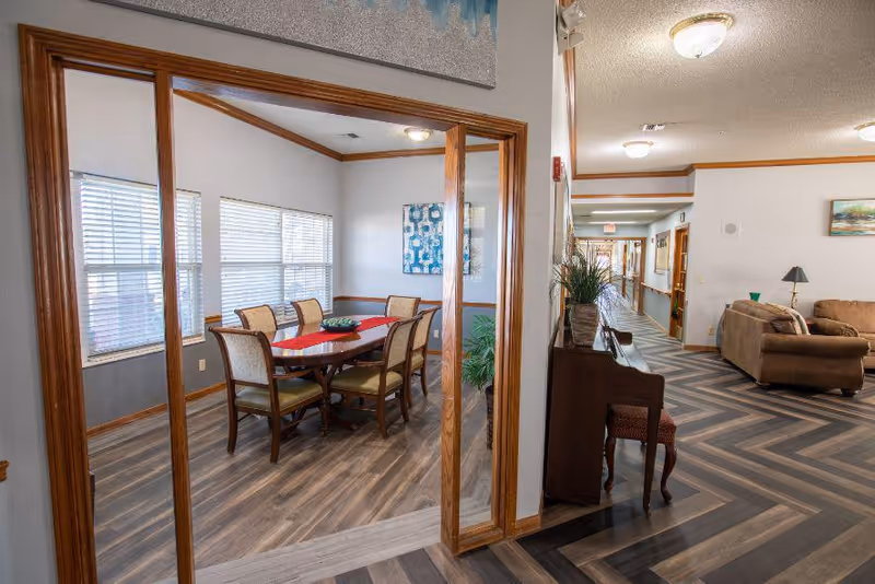 Interior view of a senior living facility showing a dining area with a wooden table and six chairs, next to a hallway with a piano and seating area with couches. The space has wood-patterned flooring, white walls with wooden trim, and multiple ceiling lights.