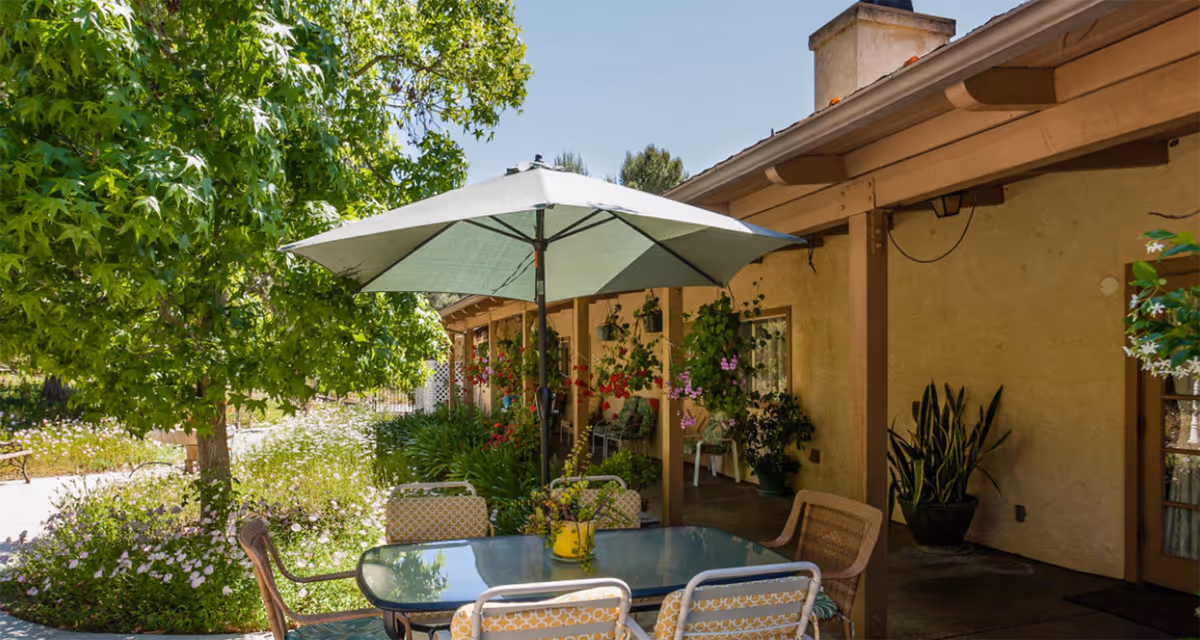 Outdoor patio area at Poway Gardens Senior Living - Mountain Vistas with a glass-top table surrounded by chairs and a large umbrella. The patio is adjacent to a building with hanging plants and greenery, and there is a tree and flowering plants nearby under a clear blue sky.