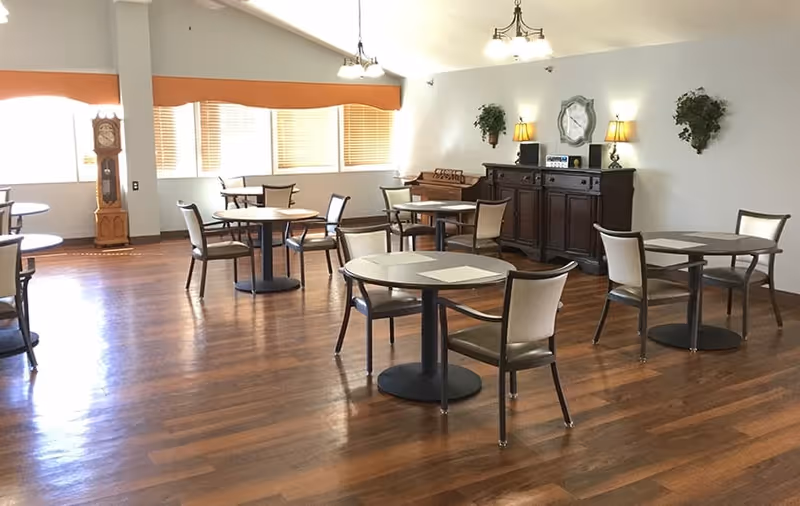 A spacious dining room with several round tables and chairs arranged on a polished wooden floor. The room has large windows with blinds allowing natural light to enter. There is a tall grandfather clock near the windows, a wooden sideboard with lamps and a clock on the wall above it, and two wall-mounted plants. The ceiling has hanging light fixtures.