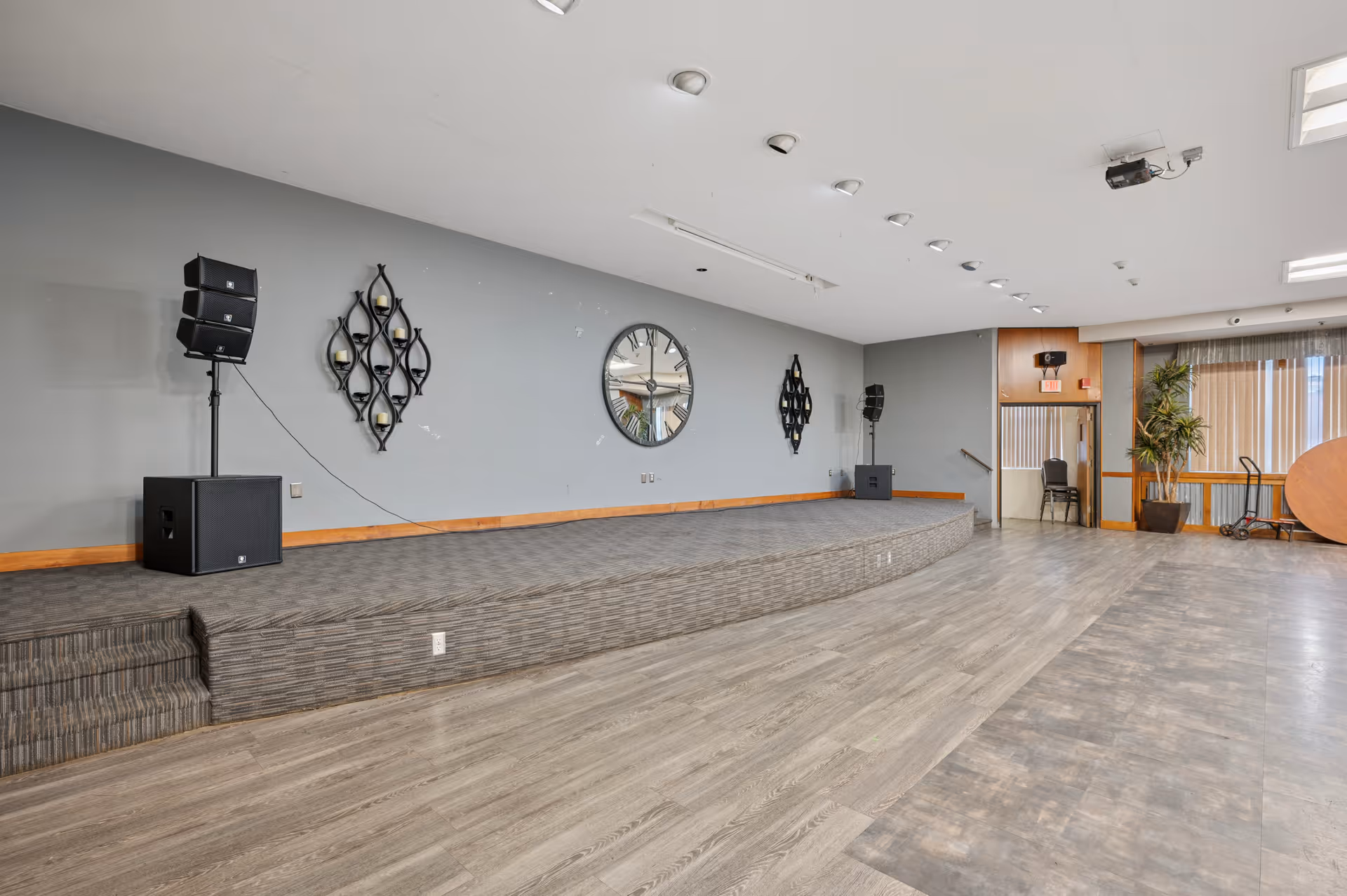 An empty event or meeting room with a raised stage area featuring speakers on stands and decorative wall hangings including a large round mirror. The room has wood-look flooring, gray walls with wooden trim, and large windows with vertical blinds. There is a potted plant and some stacked chairs near the back.