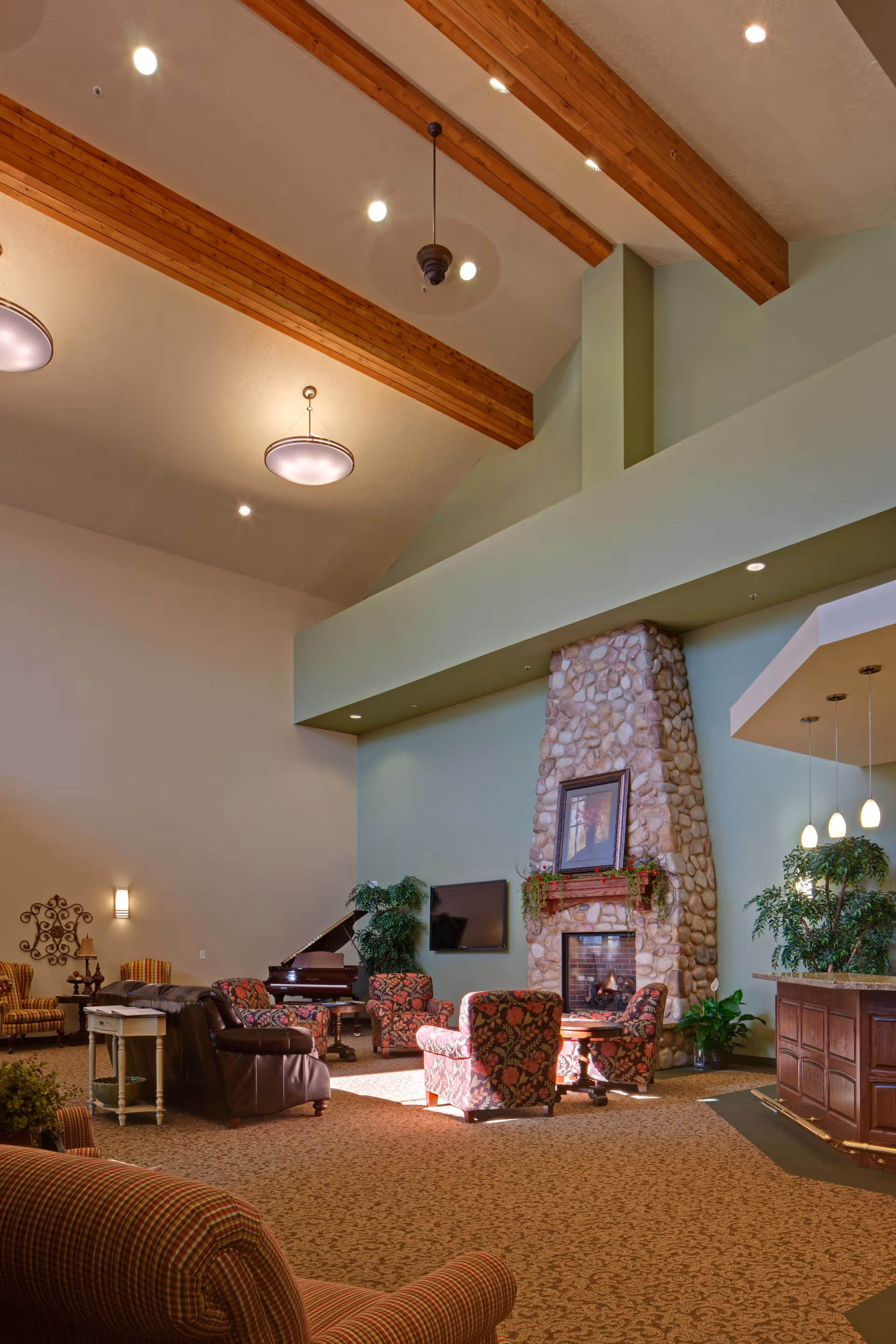 Spacious communal living room with high wooden-beamed ceiling, stone fireplace, seating area and piano.
