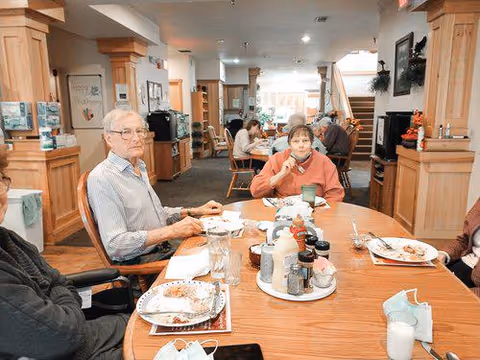 Several elderly individuals seated around a wooden dining table in a well-lit room with wooden pillars and furniture. Plates with food remnants, condiments, and glasses are on the table. The background shows more people seated and engaged in conversation or eating.
