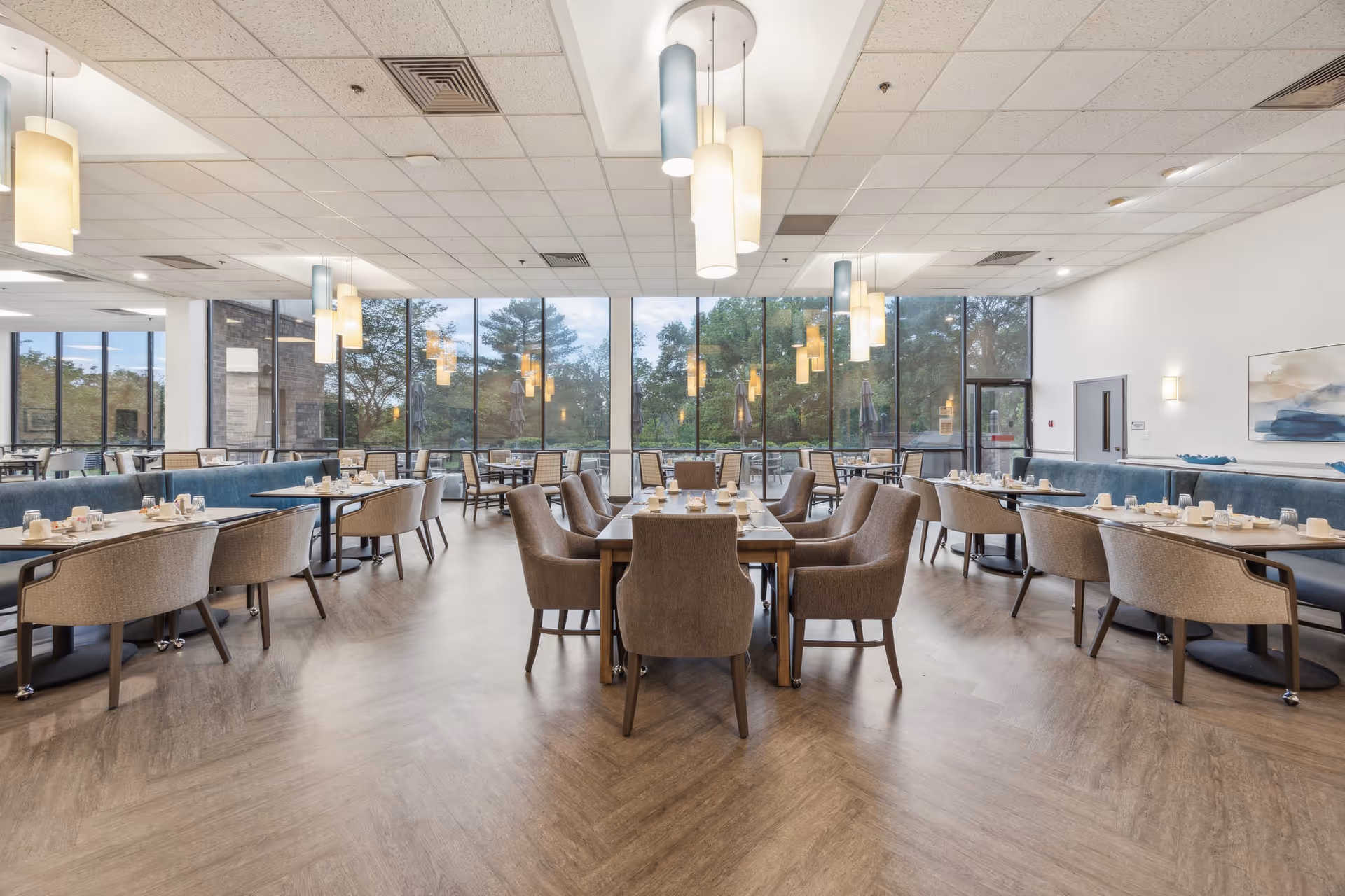 A spacious dining room with multiple tables and chairs arranged neatly. The room features large floor-to-ceiling windows providing a view of trees outside. Pendant lights hang from the ceiling, and the tables are set with cups, glasses, and napkins. The floor has a wood-like finish, and the walls are white with minimal decoration.