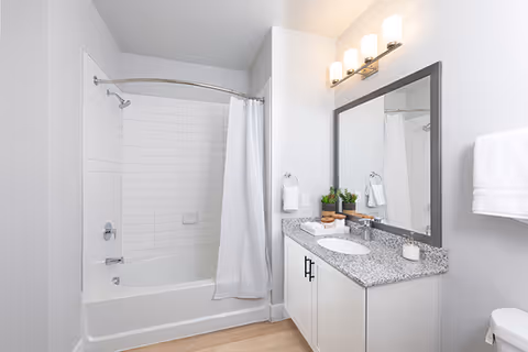 A clean and modern bathroom featuring a white bathtub with a curved shower curtain rod and white shower curtain, white tiled walls around the tub, a granite countertop with a sink, a large mirror above the sink, a wall-mounted light fixture with four lights, two small potted plants on the counter, and white towels hanging on the wall.