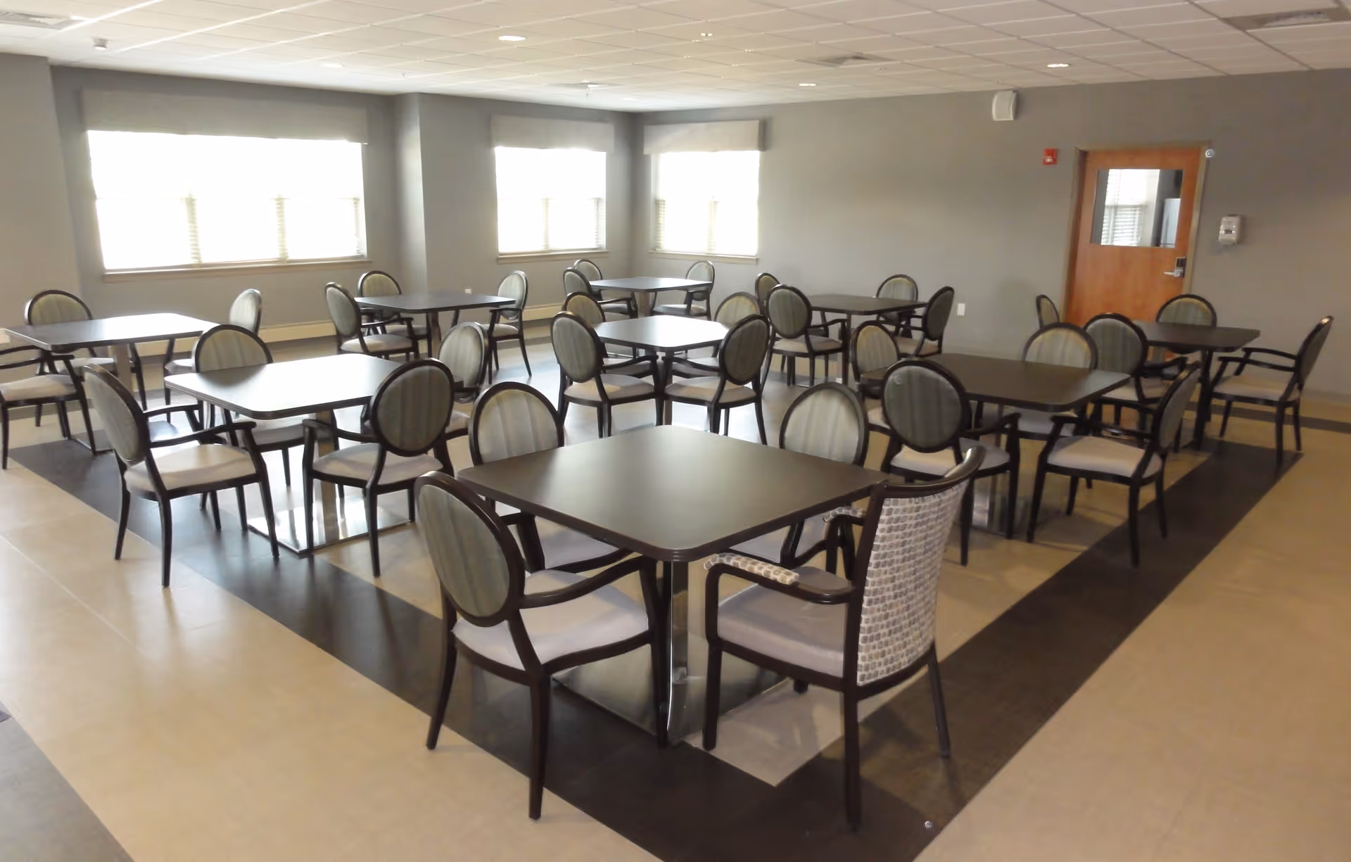 Empty dining room with multiple square tables and upholstered chairs arranged on a tiled floor.